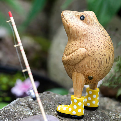 Wooden frog figurine with yellow spotted wellies posed on a rock with a fishing rod
