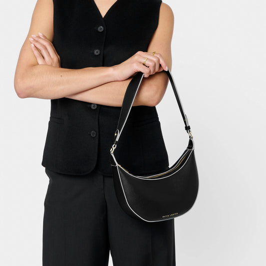 Model wearing a crescent shaped shoulder bag in black PU with contrasting sand colour wax piping