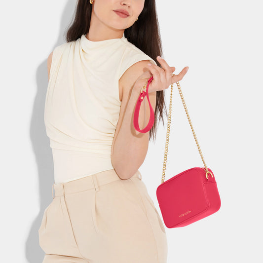 Model wearing a fuchsia camera bag with gold chain detail on the strap, textured leather and a zip top