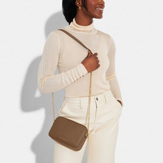 Model wearing a light brown camera bag with gold chain detail on the strap, textured leather and a zip top