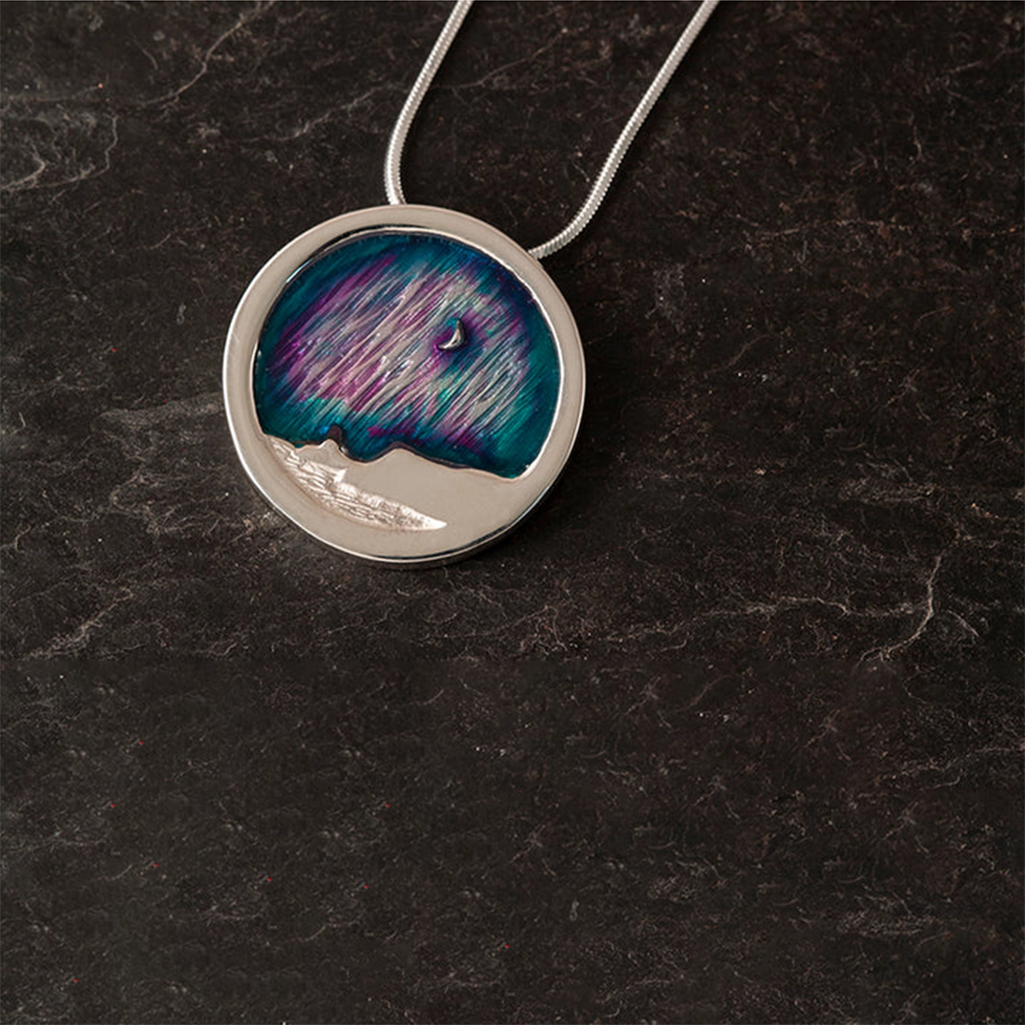 A round silver pendant with a Shetland cliff scene and purple and blue enamelled Northern Lights design
