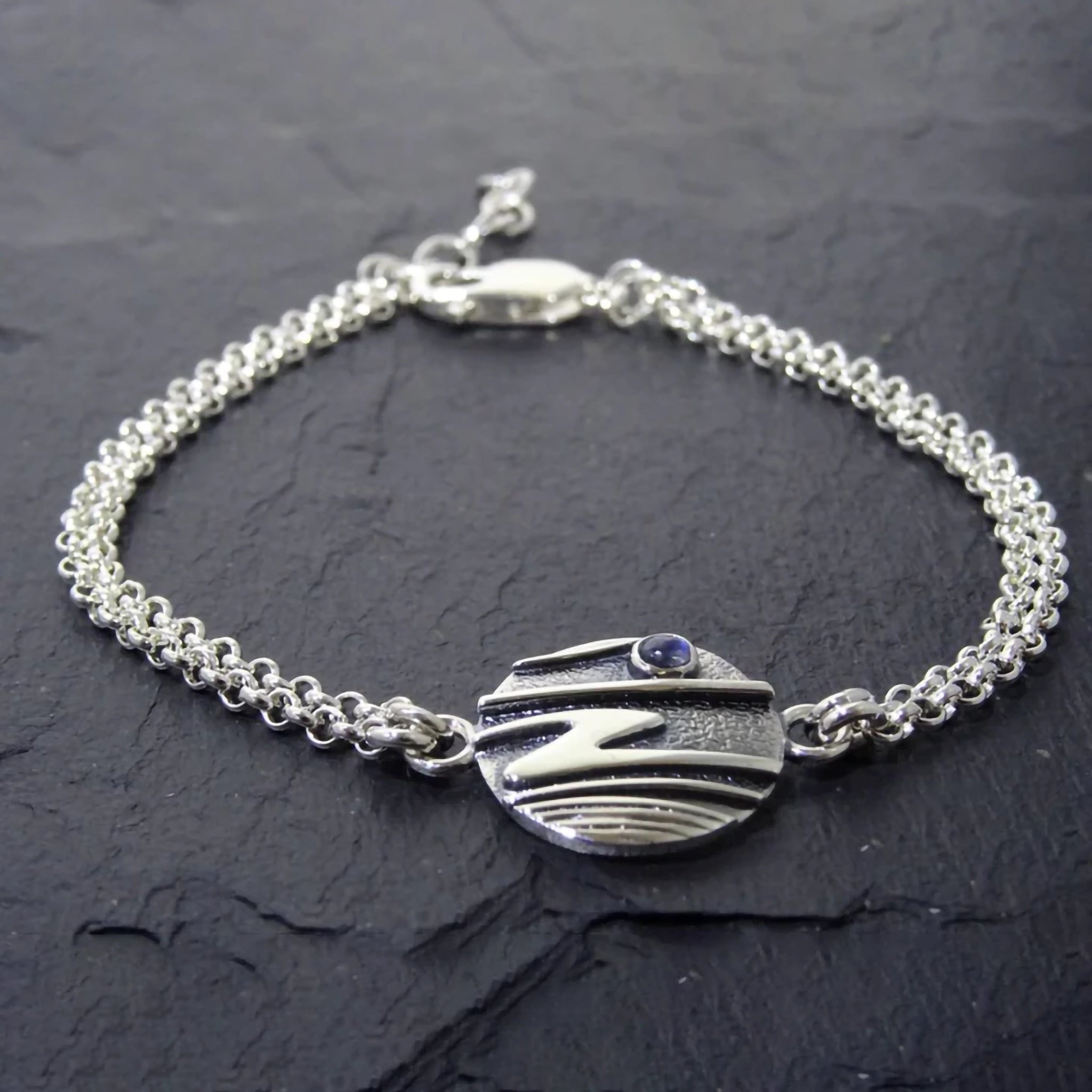 A silver double chain bracelet featuring a round pendant with raised ripple texture design and a small moonstone