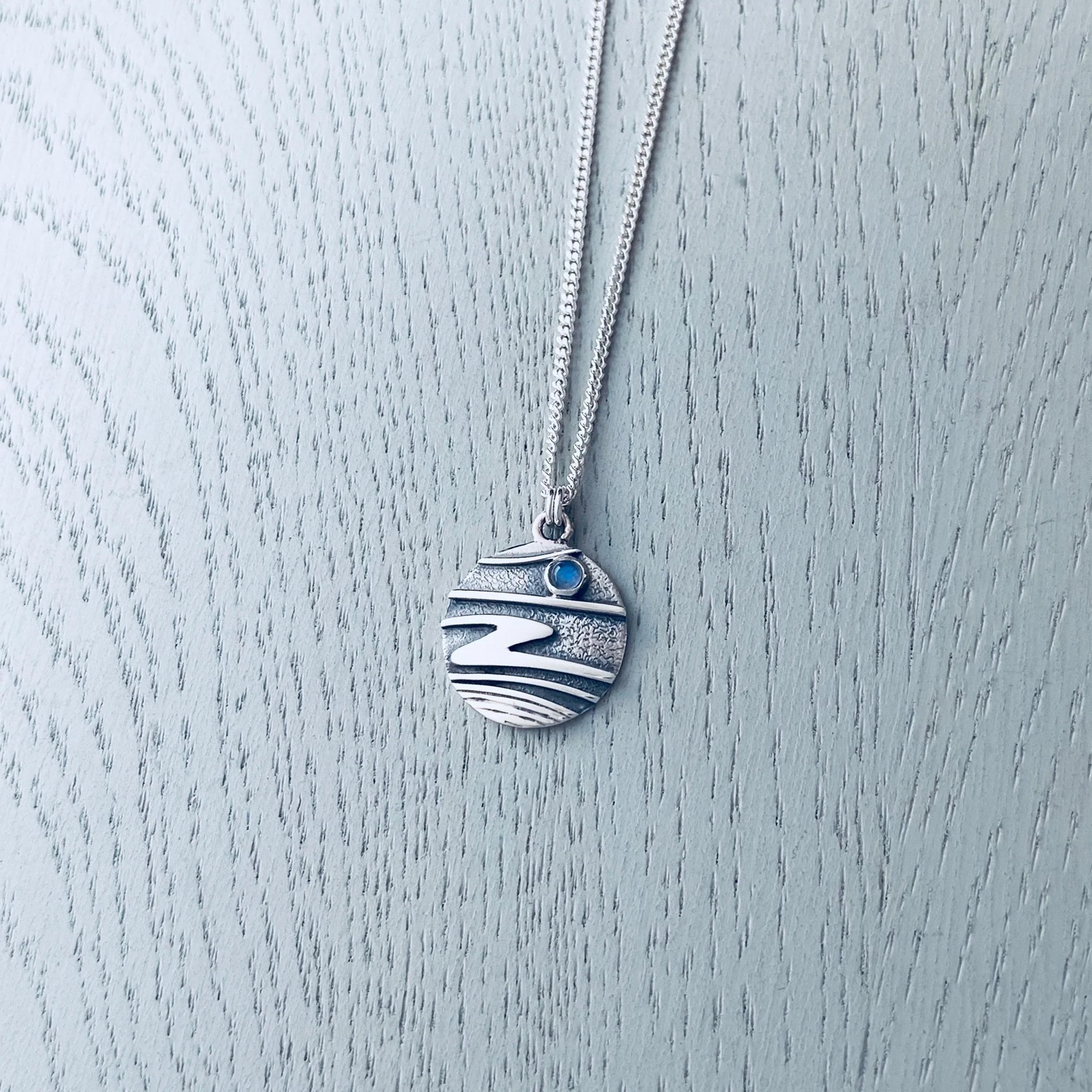 A round silver pendant on a neck wire featuring raised ripple design with dark oxidisation and a round moonstone