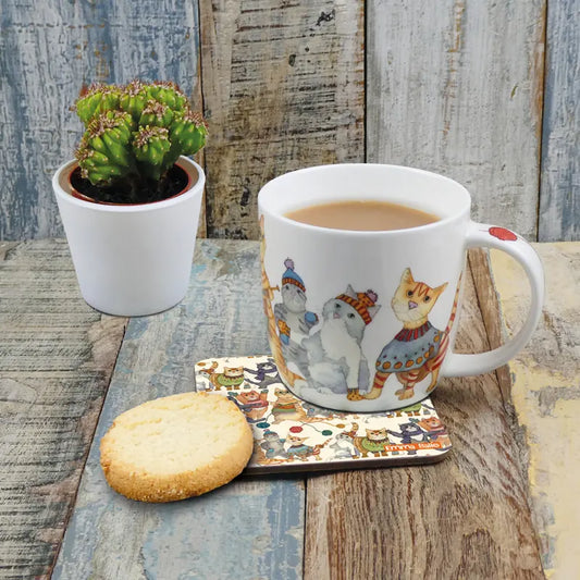 A white mug with an illustration of cats in winter clothing with tea and a biscuit