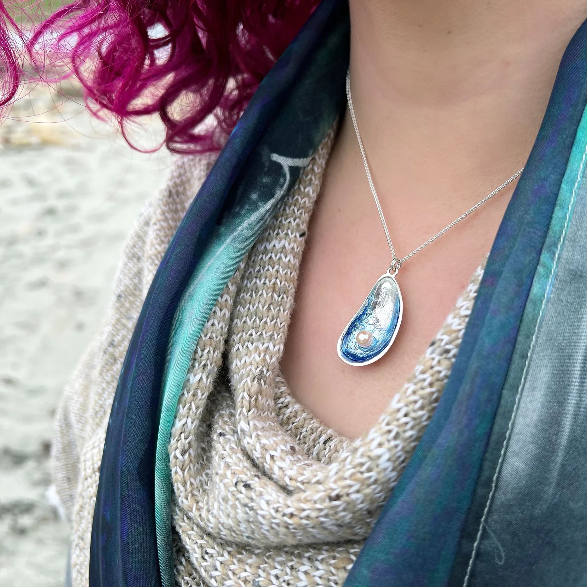 Model wearing a large silver pendant in the shape of a mussel with gradient blue enamel and a pearl on a chain