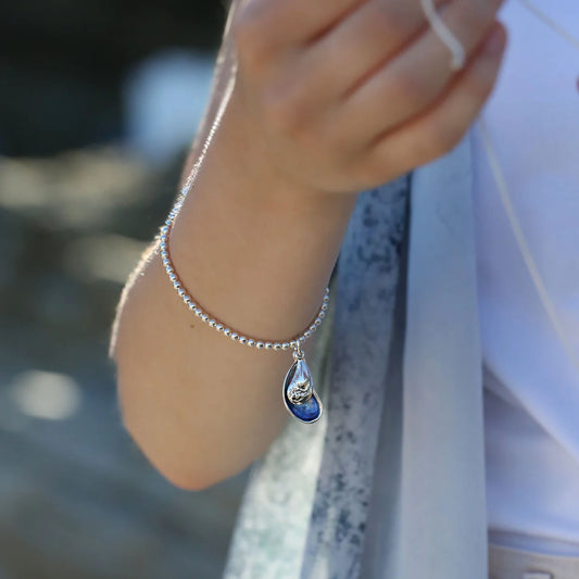 Model wearing silver beaded bracelet with a drop pendant in the shape of mussels in blue enamel