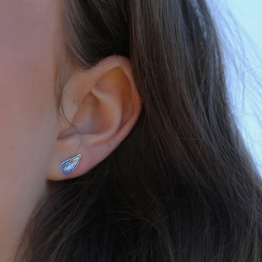 Model wearing a pair of silver stud earrings shaped like mussels with a gradient blue enamel