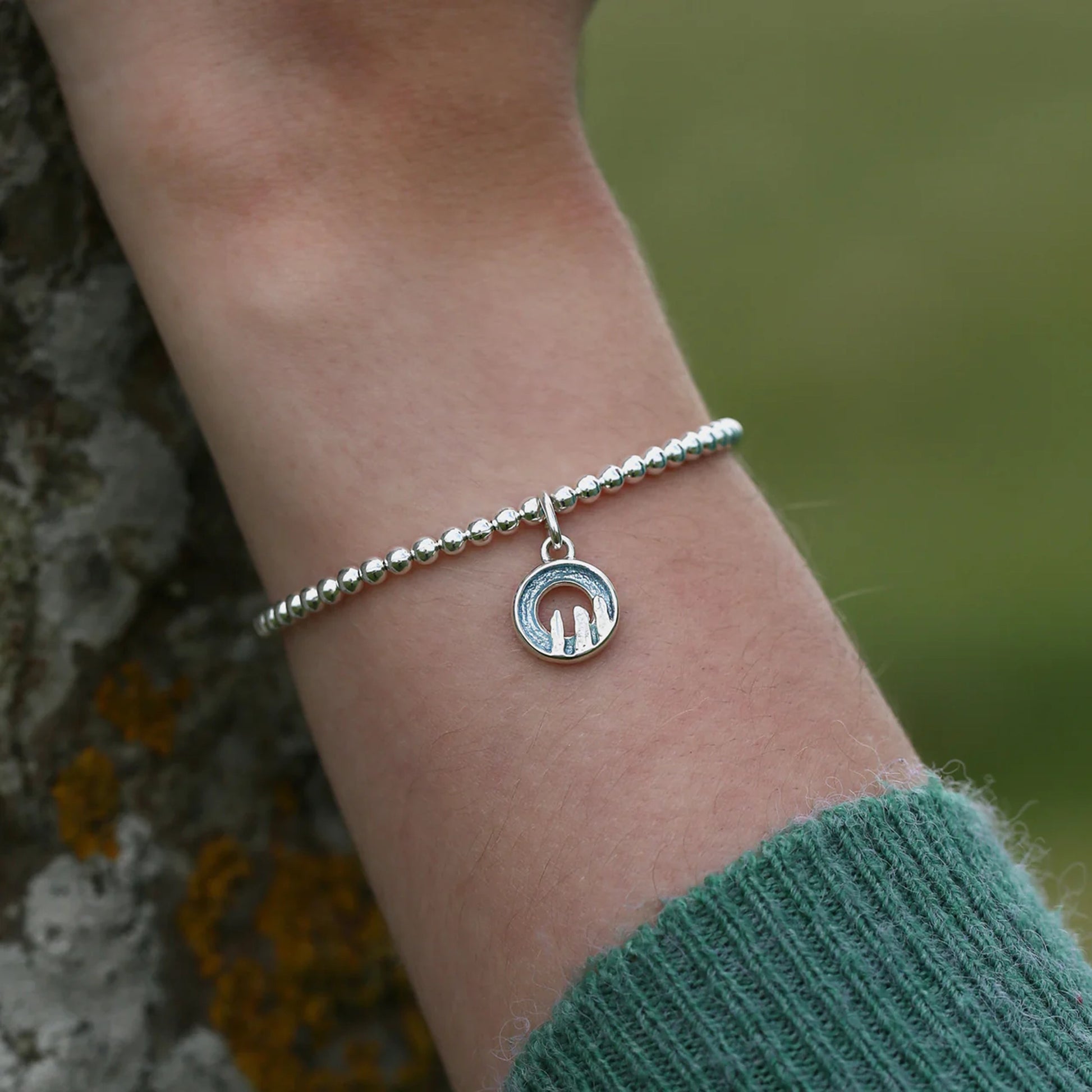 Model wearing silver stretch bracelet with a round blue grey enamelled charm with standing stone design