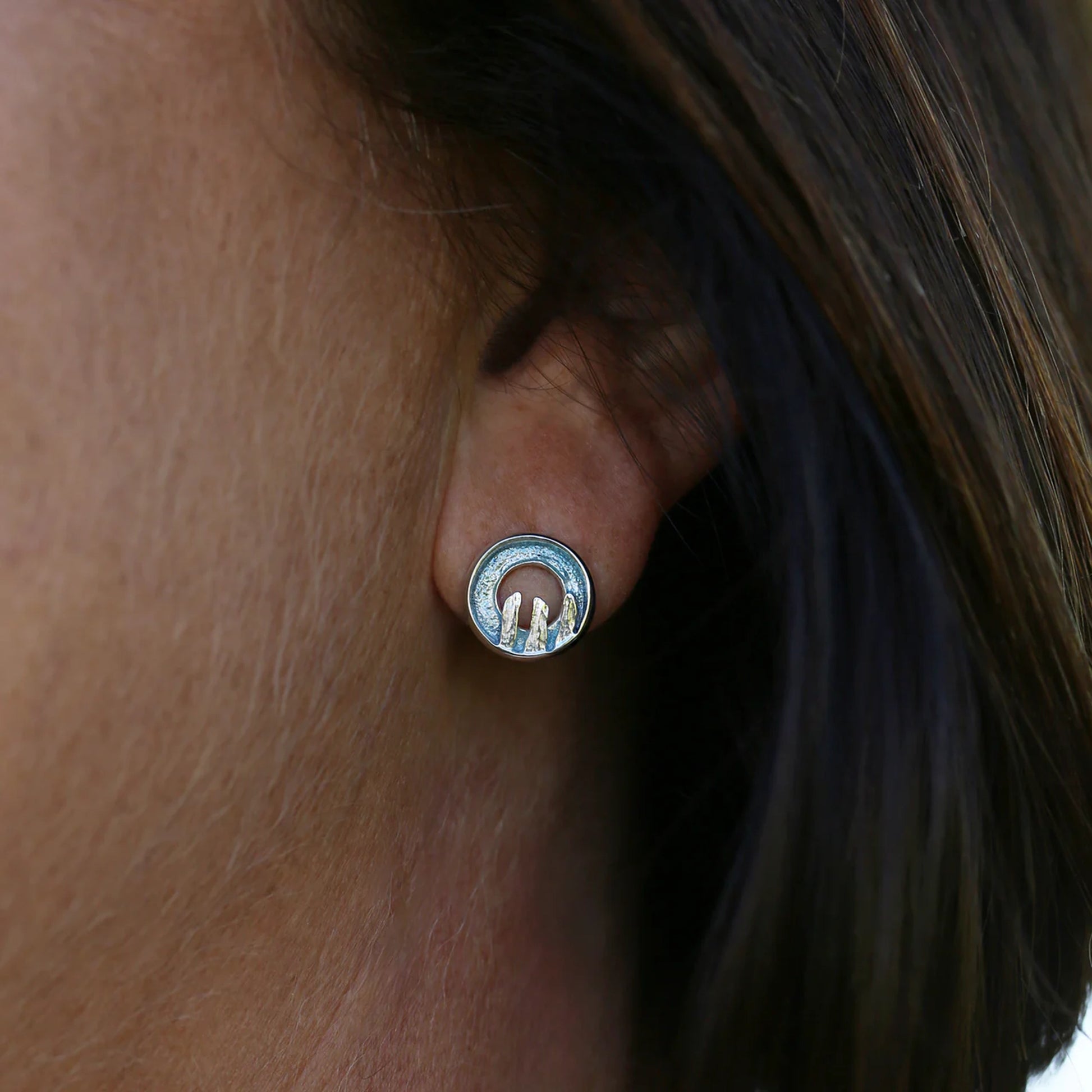 Model wearing round silver stud earrings with centre cutout, blue grey enamel, and standing stone details