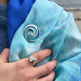 Model wearing round brooch with ocean wave design in a gradient blue enamel