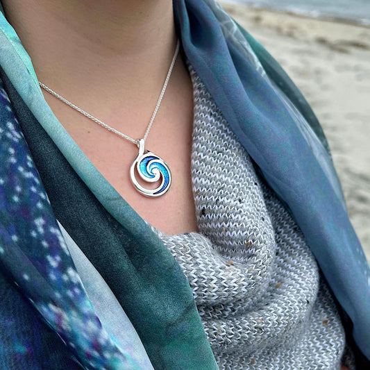 Model wearing a large round silver pendant with ocean wave design and blue enamel in the centre on a silver chain