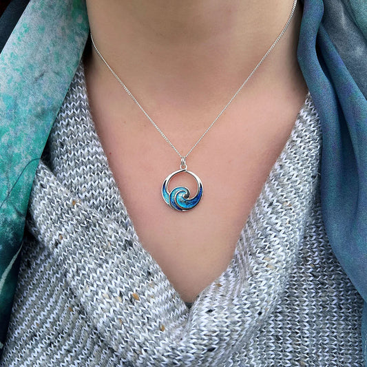 Model wearing polished silver round pendant with ocean wave design in blue enamel on silver chain