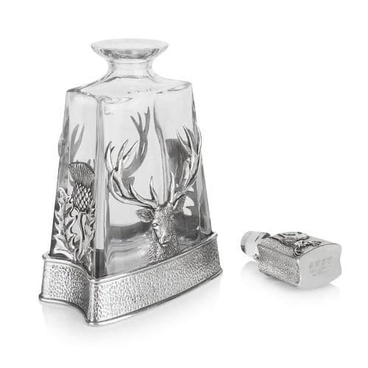 An open glass decanter with a pewter base featuring a thistle and stag head and a stopper lid featuring a standing stag