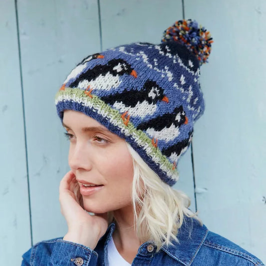 Model wearing knitted hat with colourful pompom featuring a design of a row of puffin birds in blue