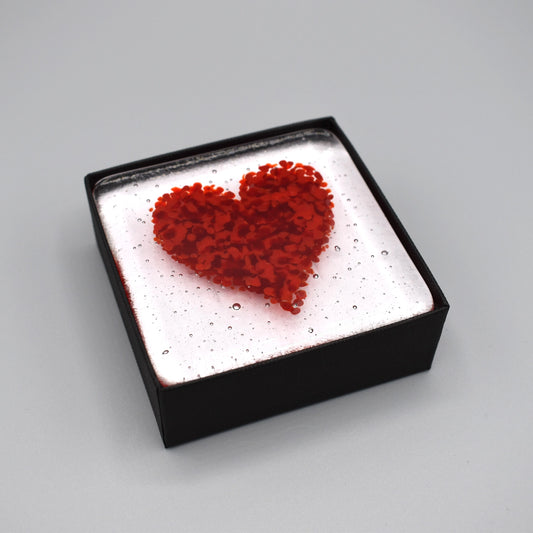 A clear glass square coaster with red heart design in a box