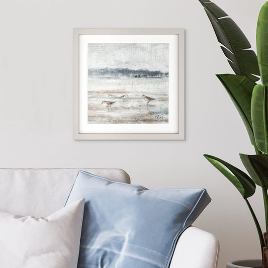 Framed print of sand pipers birds on a sandy beach at low tide in wispy greys and blues hanging in a light room.