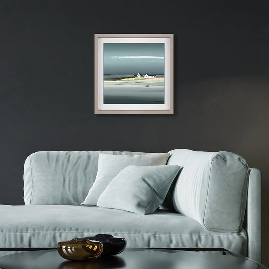 Square framed print of a seaside with blue sky, water and white cottages on a sandy shoreline hanging in a dark room.