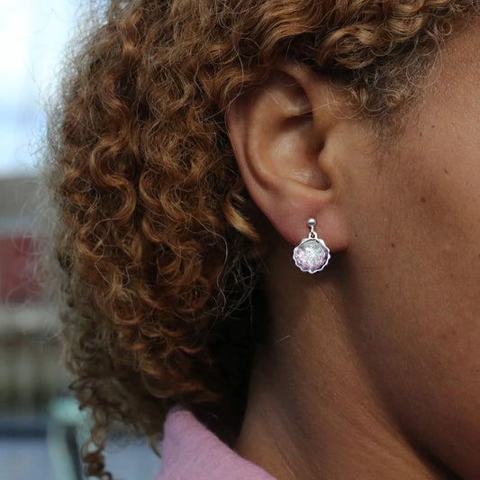Model wearing a pair of small silver drop earrings shaped like scallop shells with gradated pink enamel centres