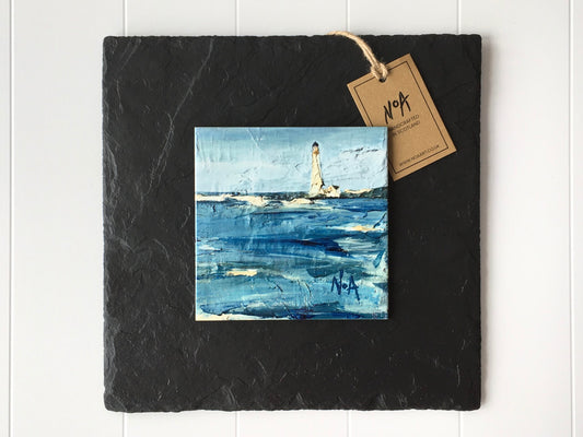 Wall art of a lighthouse and sea in vibrant blues done in an abstract style onto a square ceramic tile mounted on square slate