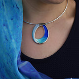 Model wearing large silver necklet with bright blue enamel in a simple abstract ocean wave shape on a silver neck wire