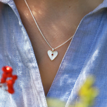 Model wearing heart shaped silver pendant in white enamel with a red garnet stone in the centre and a silver chain