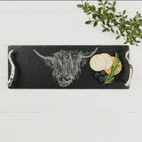 A long and thin slate serving tray with an engraved Highland cow and silver horn shaped handles staged on a table