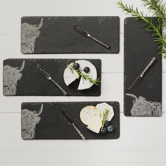 A staged set of four slate cheeseboards with engraved Highland cow and a steel cheese knife on each
