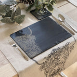 A slate placemat with engraved Highland cow staged on a table with matching tablecloth and coaster