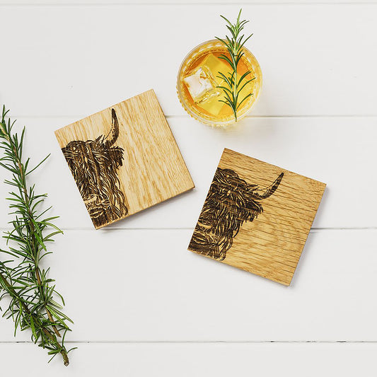 Two square wooden coasters with an engraved Highland cow on each staged on a table with a glass of whisky