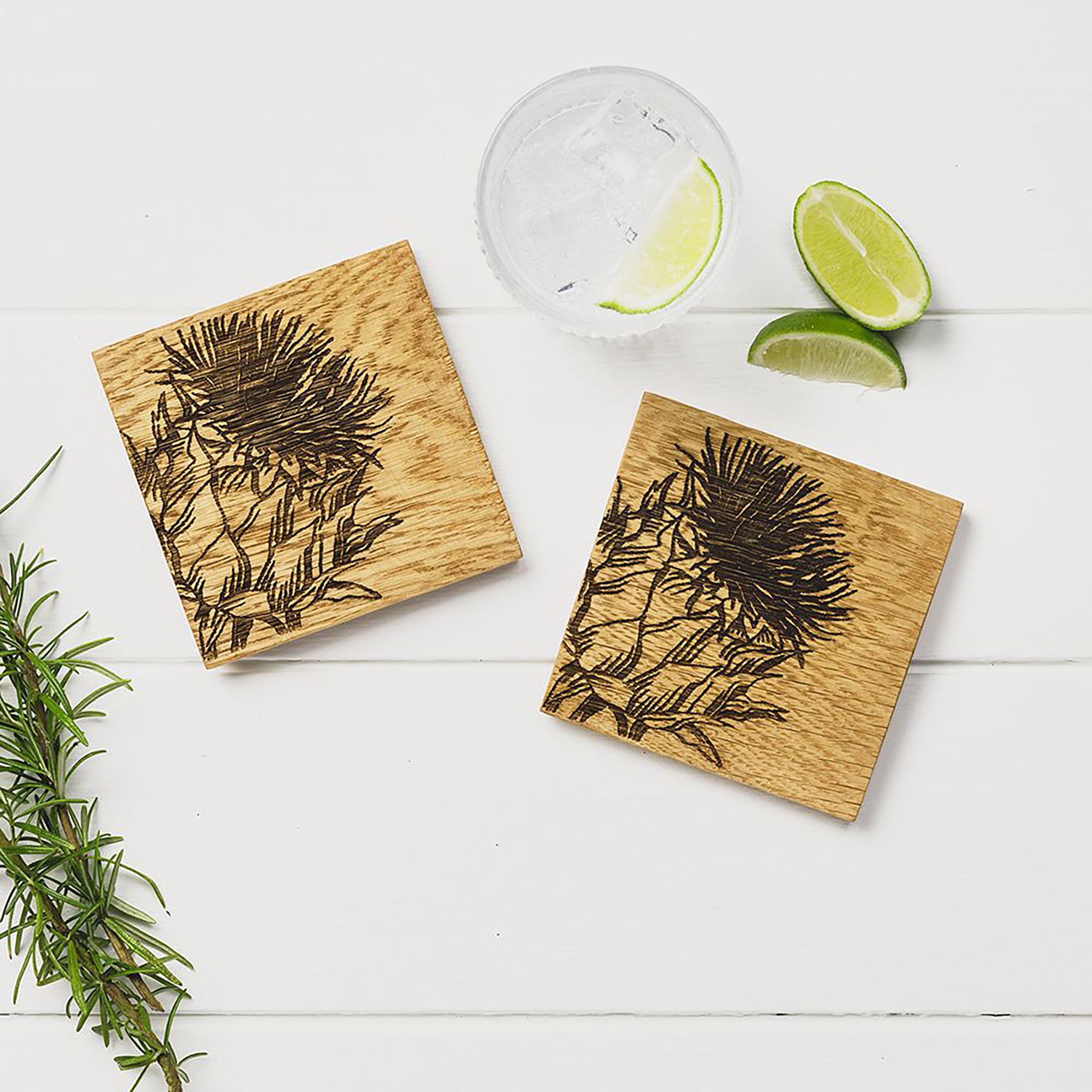 Two square wooden coasters with an engraved Scottish thistle on each staged on a table with a glass of gin