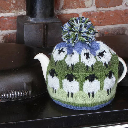 A knitted tea cosy featuring sheep in field in green and blue lifestyle