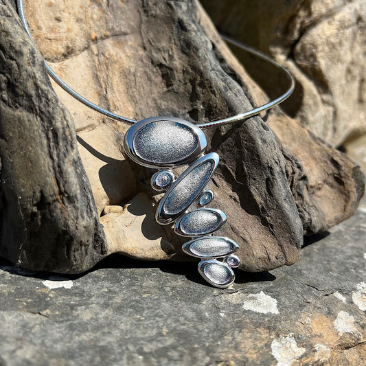 Silver necklet with organic stacked pebble shaped pendant in pearl grey enamel with silver neck wire posed outside