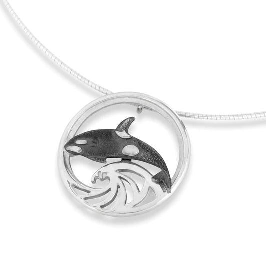 A round silver pendant featuring an oxidised orca on a wave design on a silver neck wire