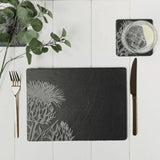 Two slate placemats with engraved Scottish thistle staged on a table with gold cutlery