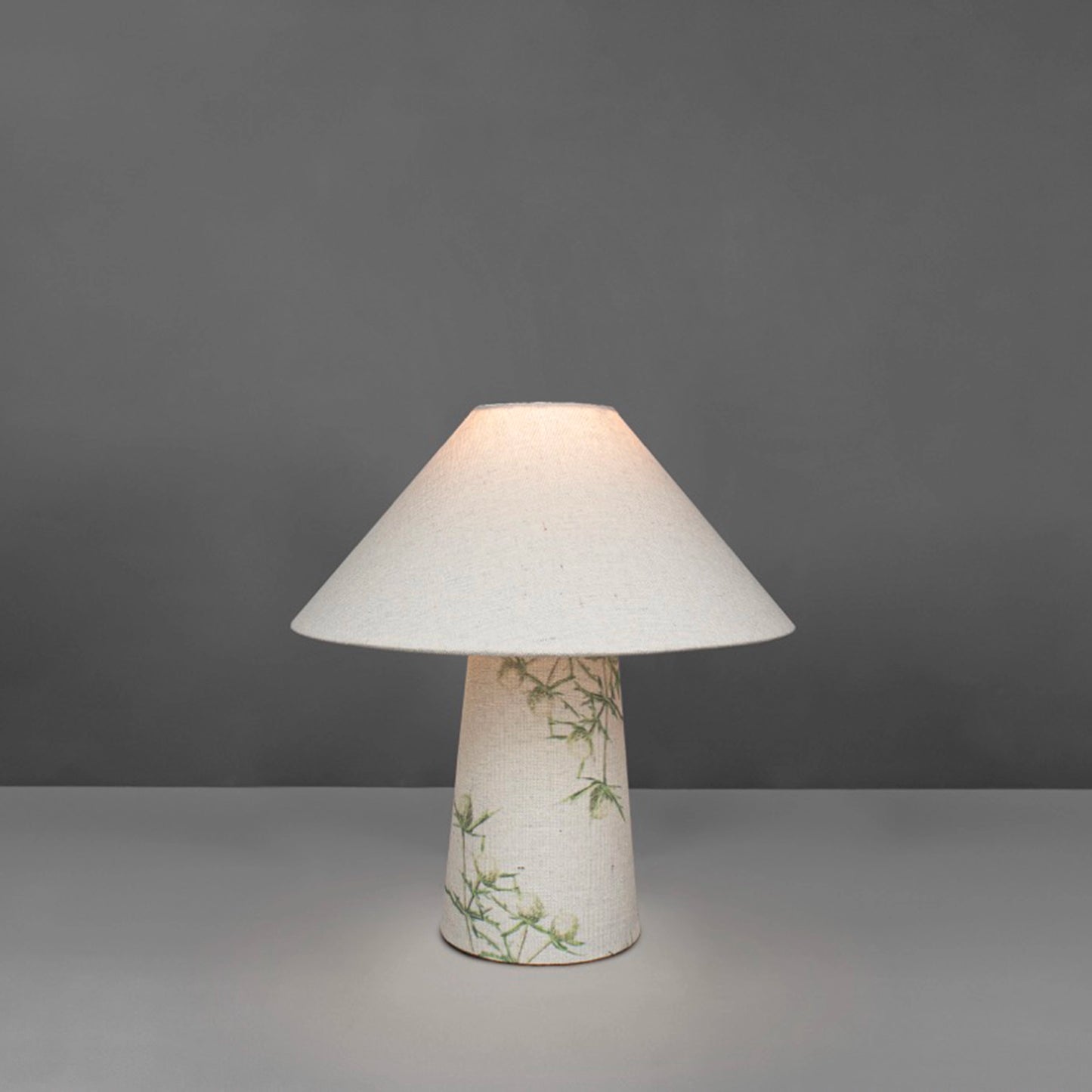 A small fabric table lamp with shade and a thistle design on the conical base lit up