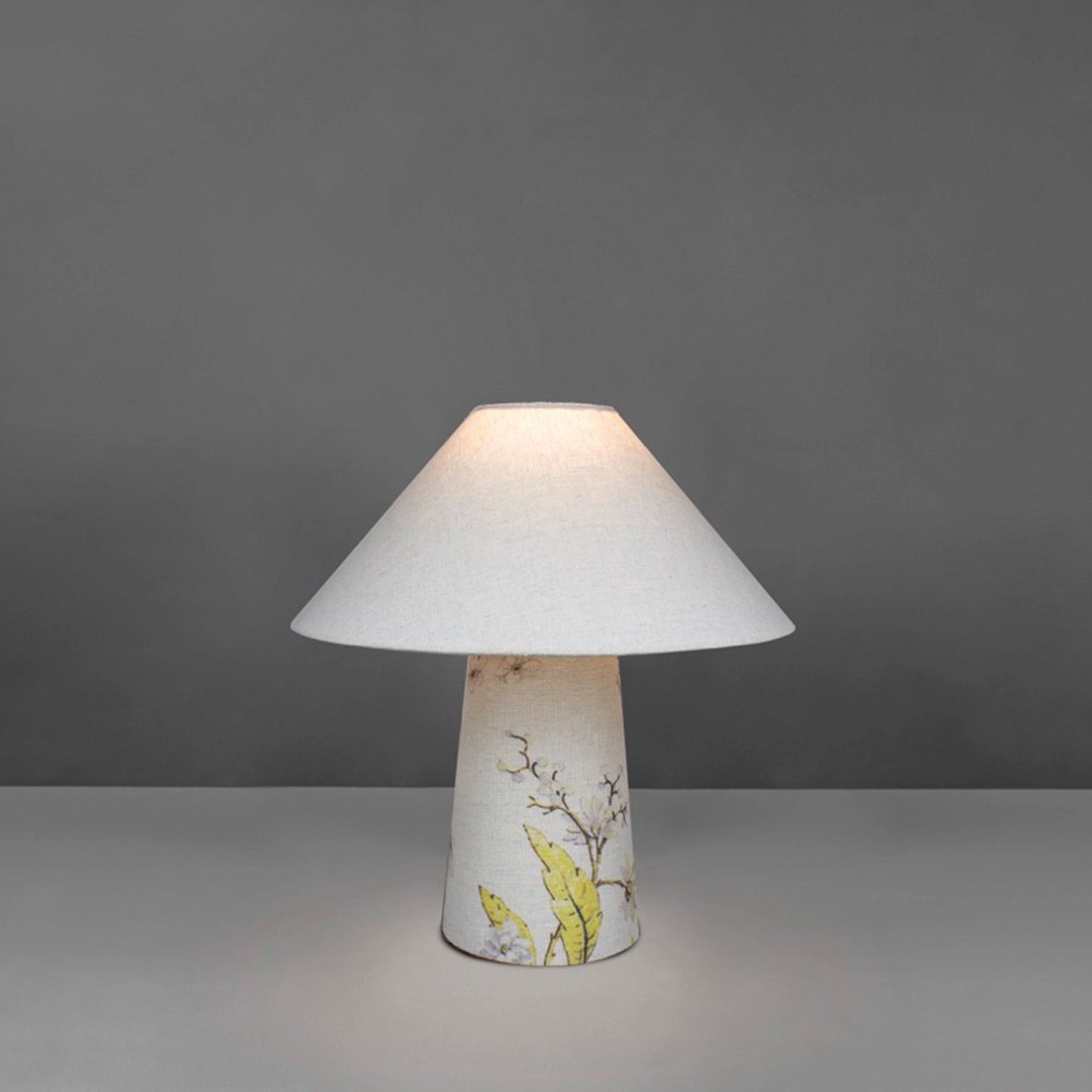 A small fabric table lamp with shade and a yellow blossom design on the conical base lit up