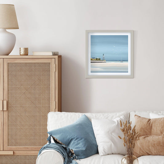 Square framed print of a seaside with blue sky and water, boats on the horizon and a lighthouse hanging in a light room.
