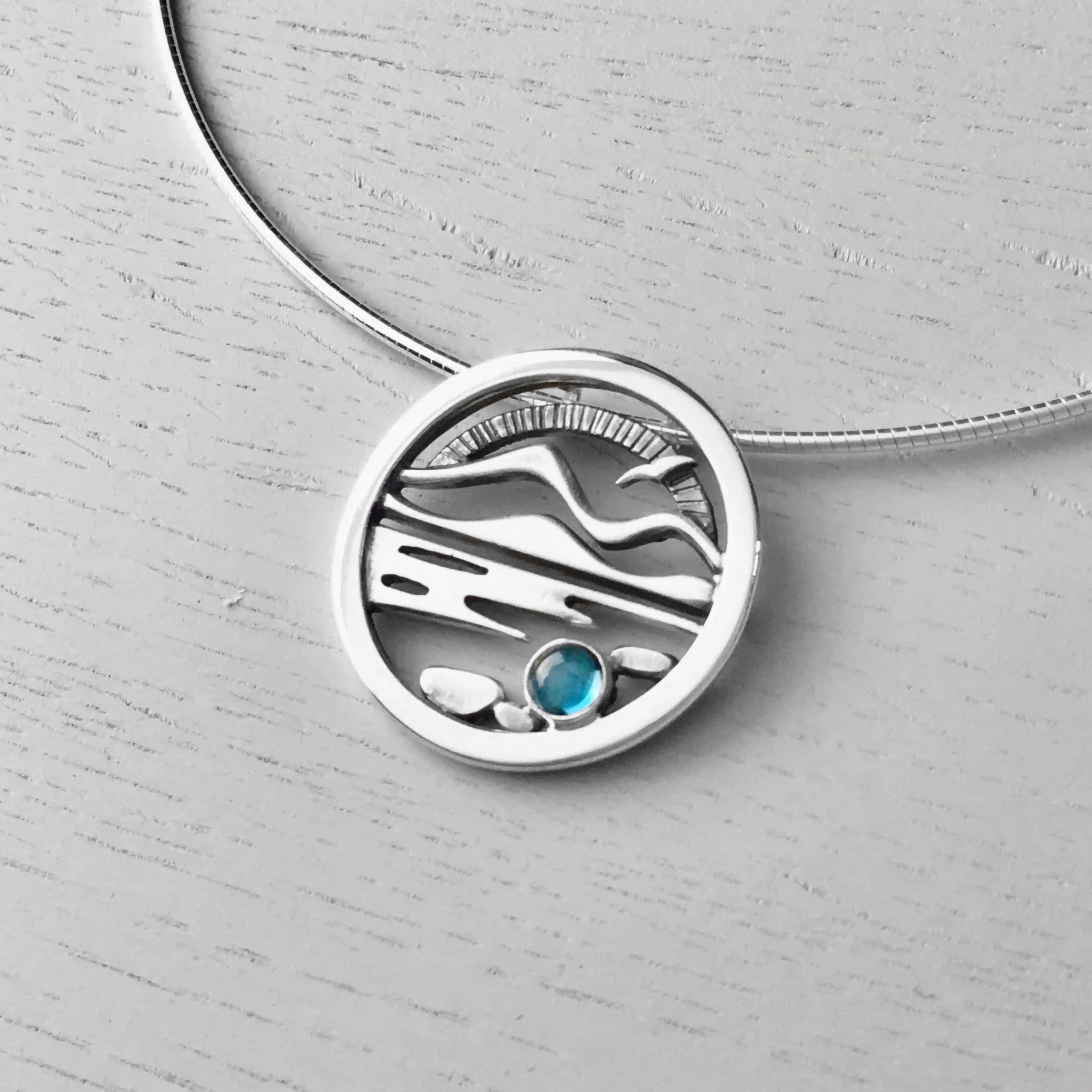 A round necklet featuring a Highland scene with water, Munros, a bird and a blue topaz stone