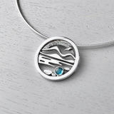A round necklet featuring a Highland scene with water, Munros, a bird and a blue topaz stone