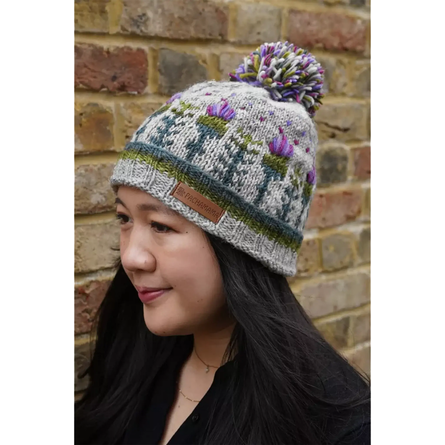 Model wearing knitted grey hat featuring a repeating thistle pattern with hearts and a pompom