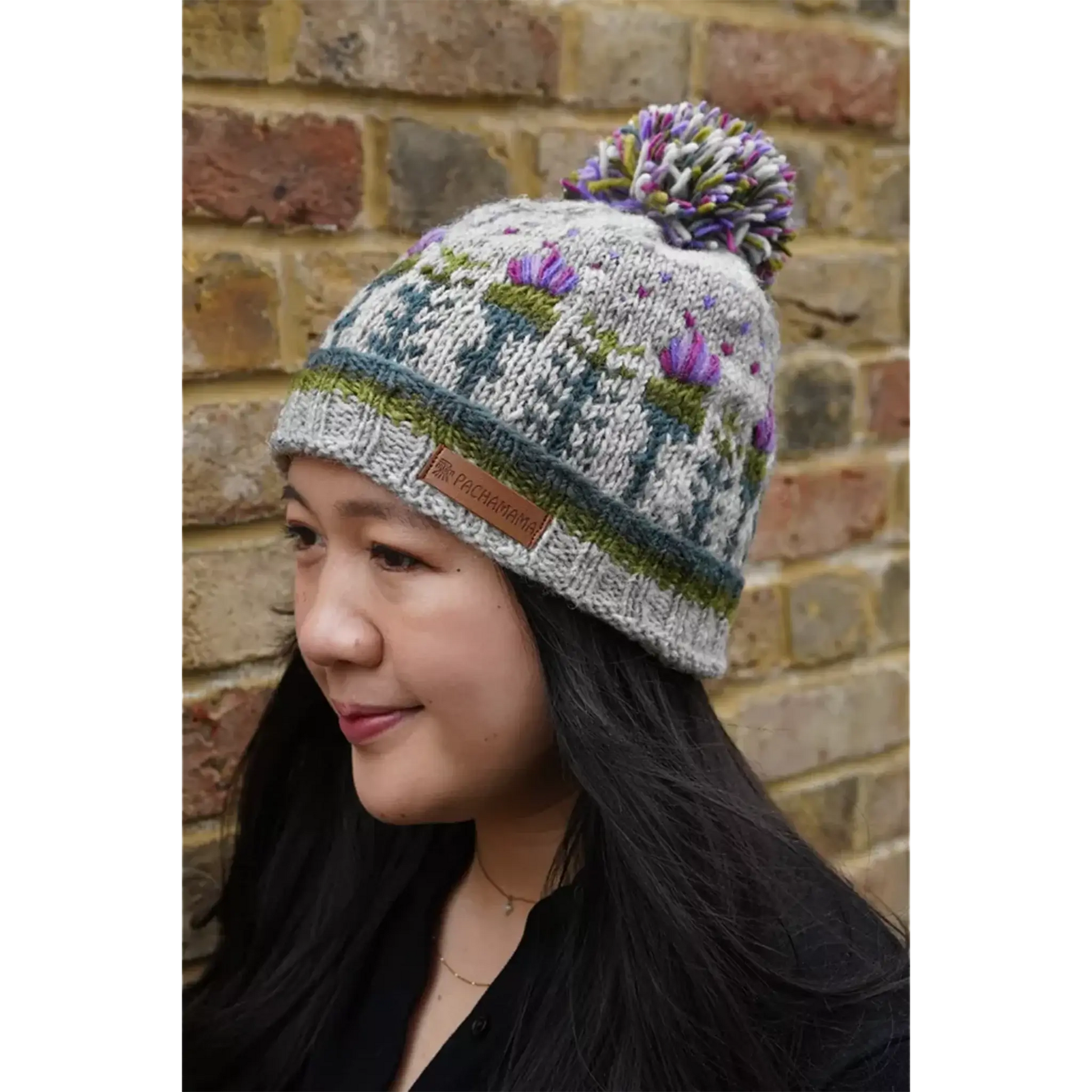 Model wearing knitted grey hat featuring a repeating thistle pattern with hearts and a pompom