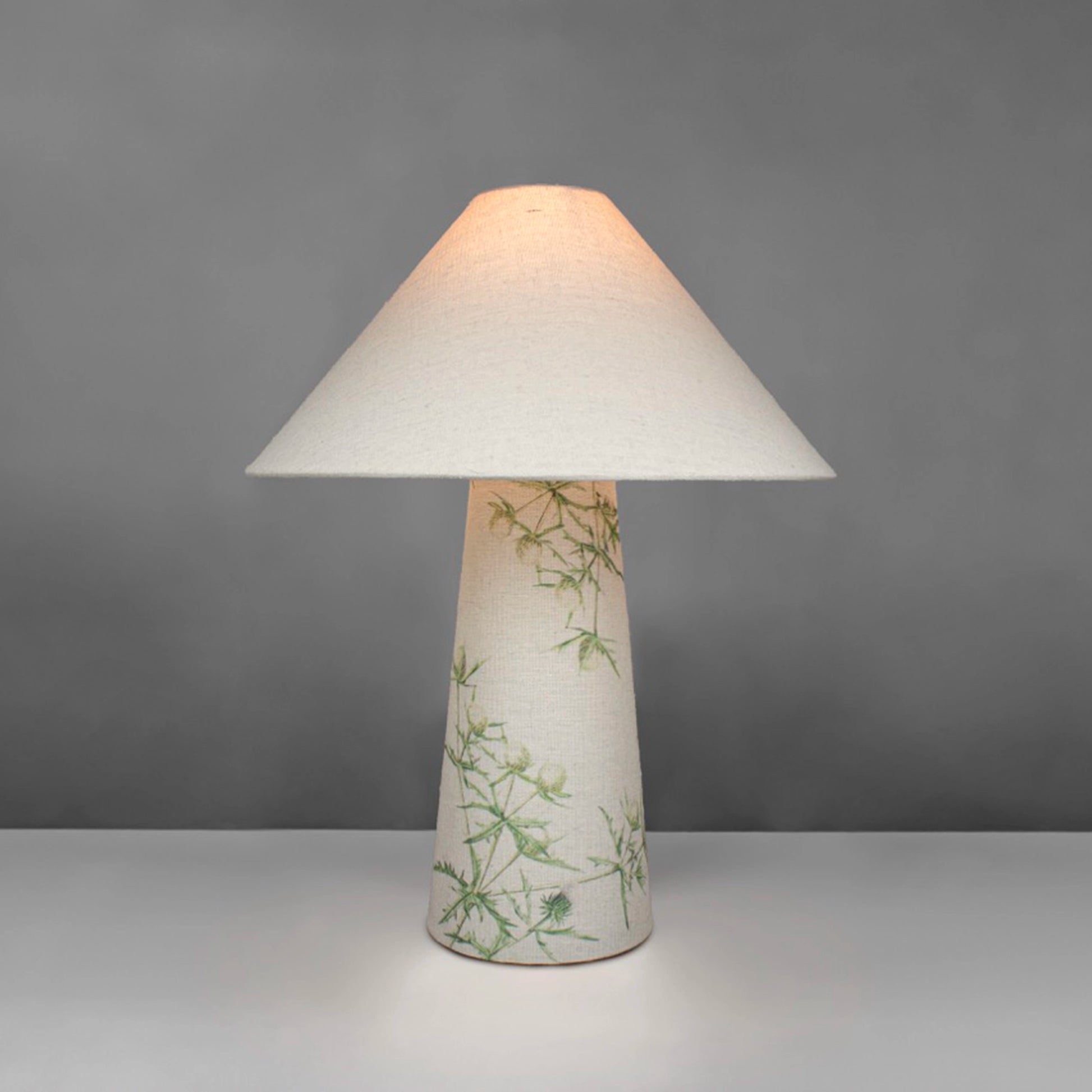 A fabric table lamp with shade and a thistle design on the conical base - lit up