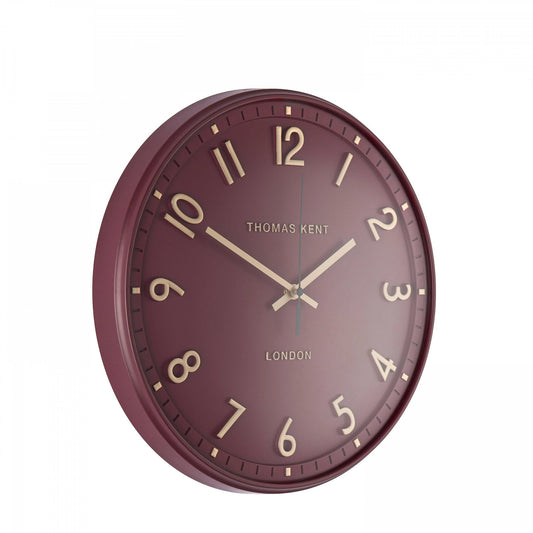 A simple and modern round wall clock in a berry colour with gold hands and numbers side view