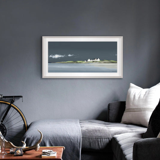 Panoramic framed print of a seaside with blue sky and water and white cottages on a green island hanging in dark room.