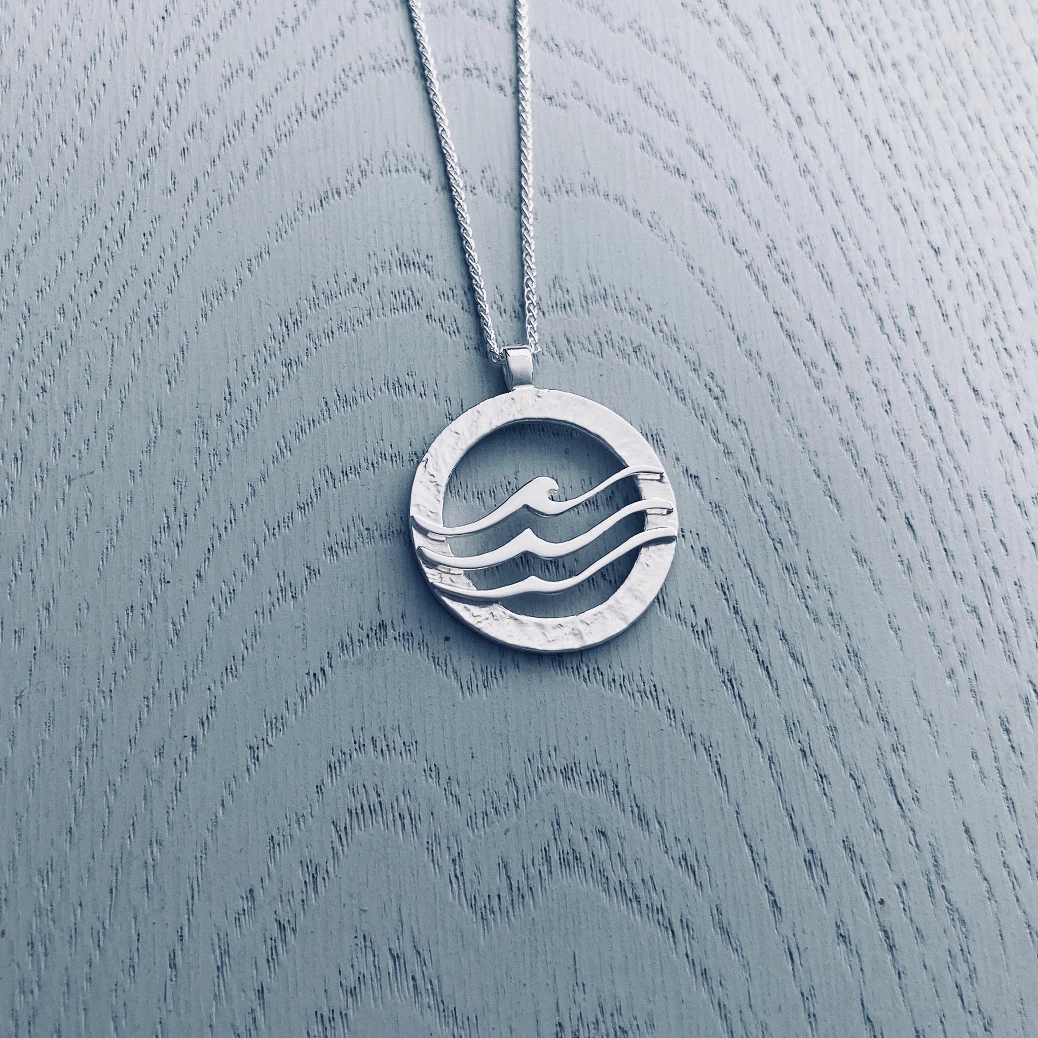 Large round silver pendant with textured frame overlaid with three waves 