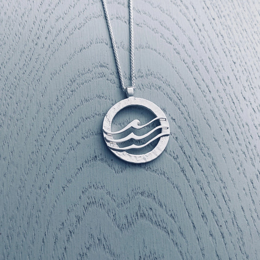 Large round silver pendant with textured frame overlaid with three waves 