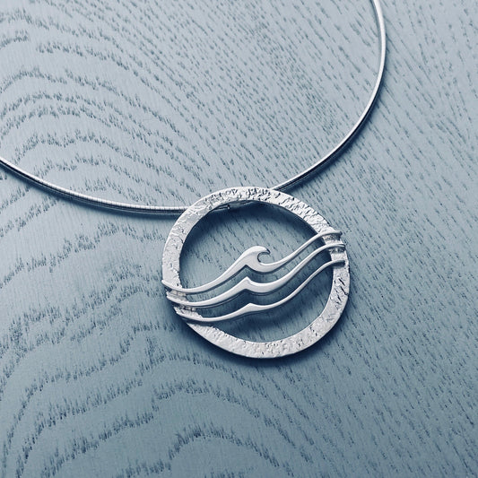 A round silver pendant on neck wire featuring a textured frame overlaid with three waves