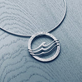 A round silver pendant on neck wire featuring a textured frame overlaid with three waves