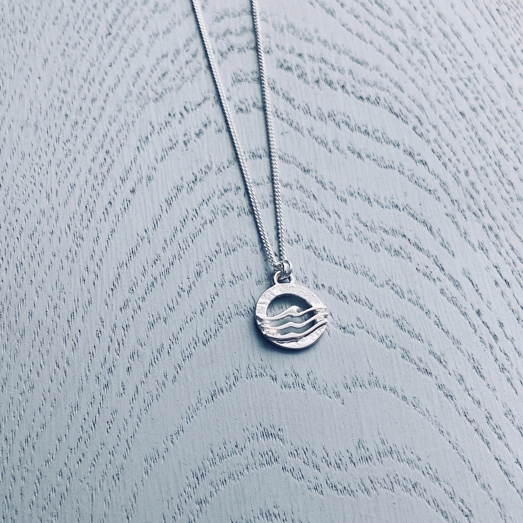 A round silver pendant on neck wire featuring a textured frame overlaid with three waves