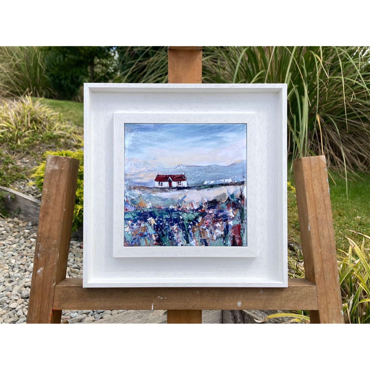 A ceramic tile printed with an abstract painting of a cottage and flowers mounted on a white frame